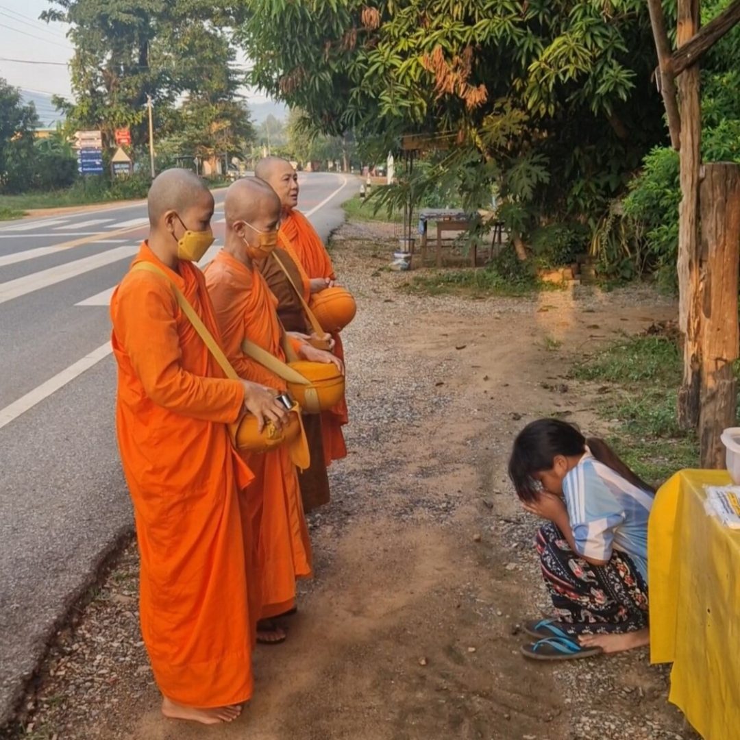 Tre monache buddiste in abiti arancioni ricevono cibo da una bambina durante un rituale di elemosina in Thailandia
